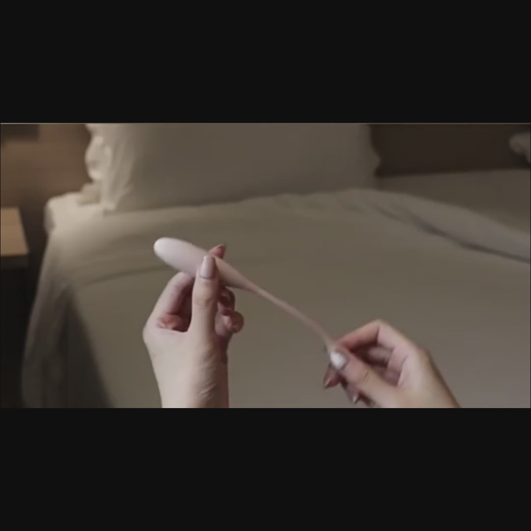 Pink mini wearable vibrator held beside a finger on a bed.